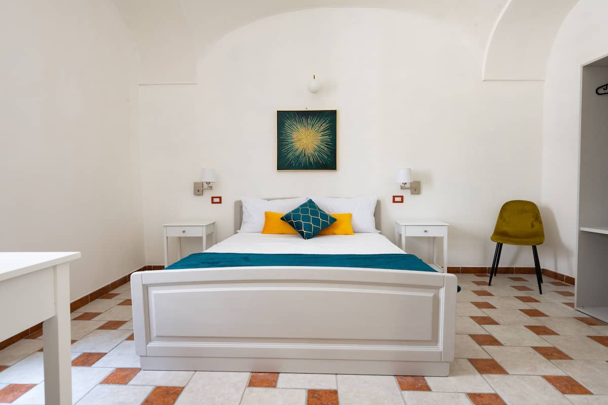 Een slaapkamer met een wit bed, twee nachtkastjes, een stoel en een wandlamp.