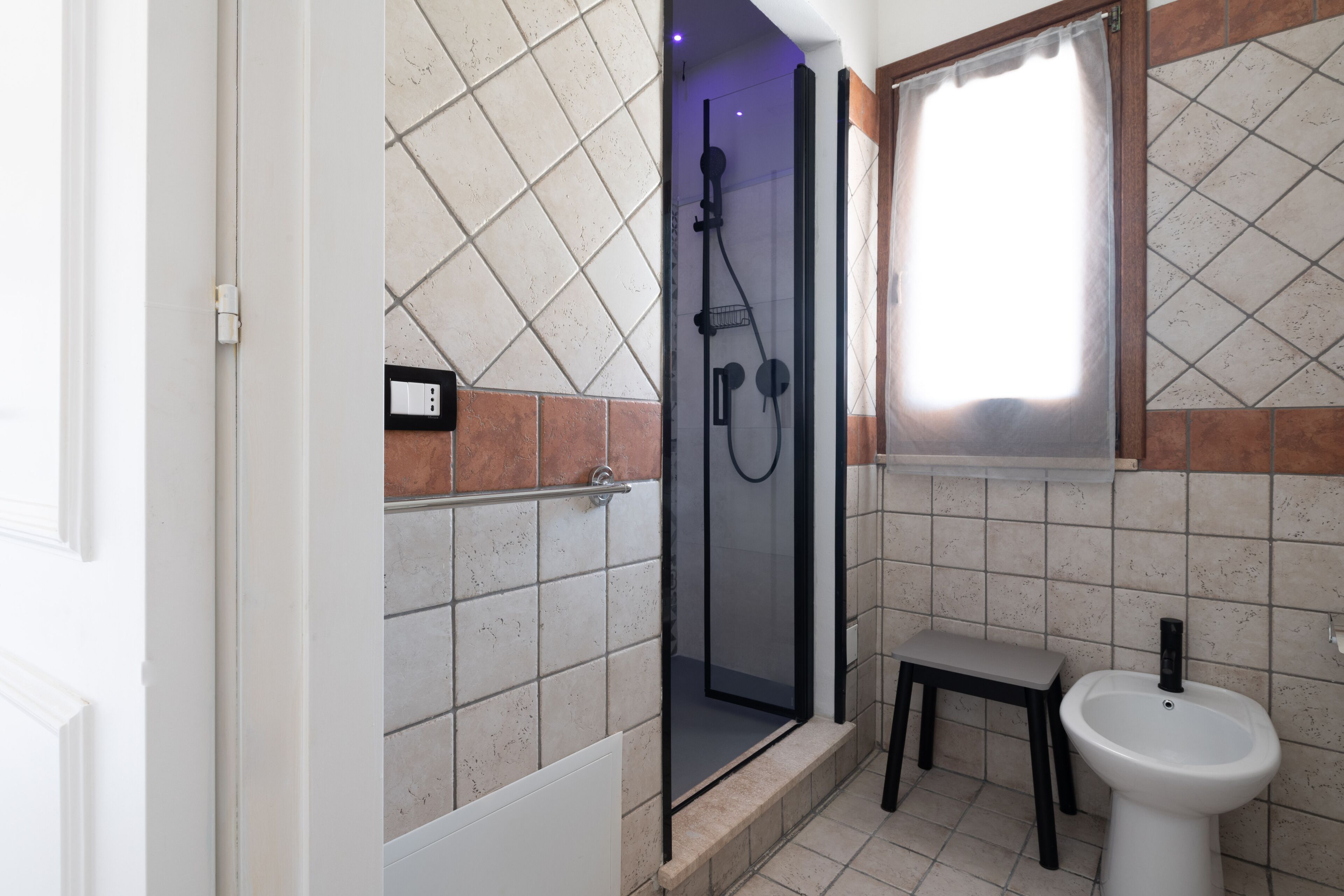 A bathroom with a glass-enclosed shower, a small stool, and a pedestal sink.