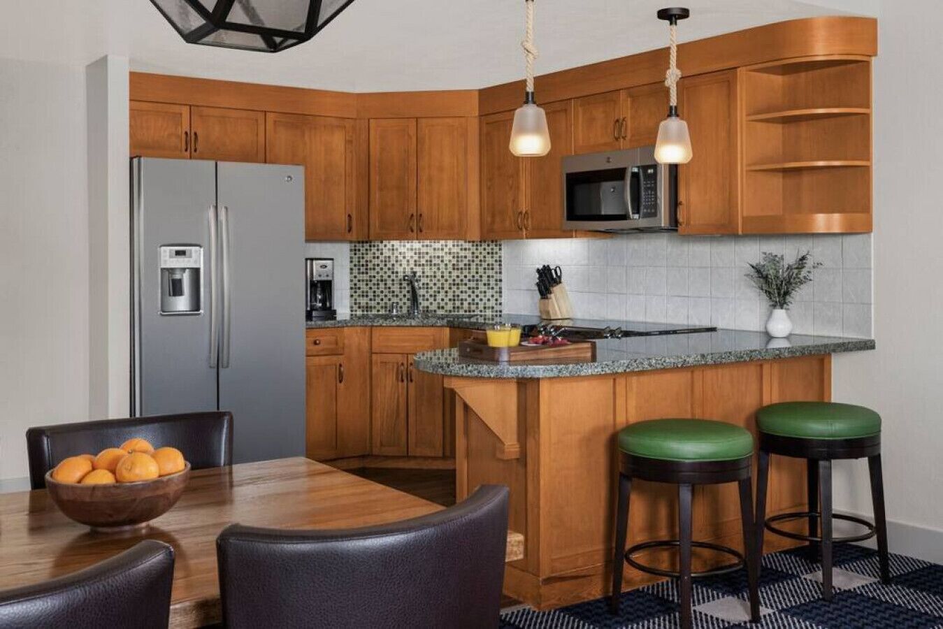 Une cuisine moderne avec des meubles en bois, un réfrigérateur en acier inoxydable, un four à micro-ondes et un plan de travail en granit. On y trouve une table à manger avec des chaises et un saladier rempli de fruits.