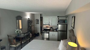 A compact hotel room with a bed, bedside lamp, kitchenette with a stove and refrigerator, a small dining area with chairs, and a mirror. - Oceanfront Efficiency T508 (Myrtle Beach)