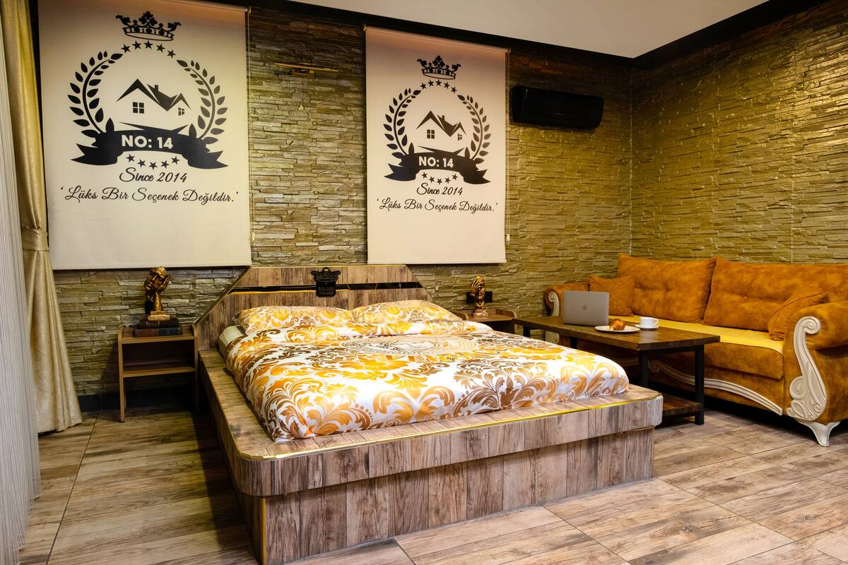 A modern hotel room with a wooden bed, a sofa, and a laptop on a wooden table. There are two framed awards on the wall.