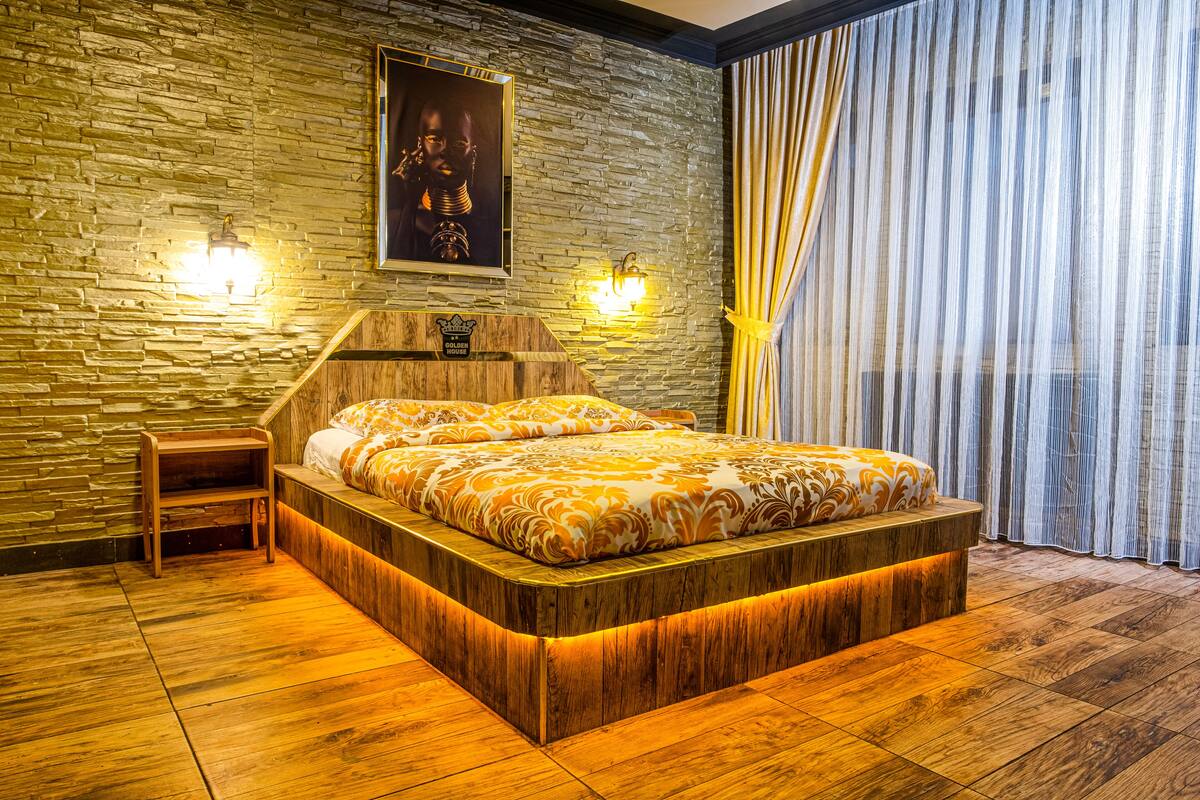 A bedroom with a stone wall, a bed with a wooden headboard, a nightstand, and a large window with curtains.