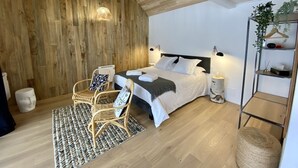 Una camera da letto moderna con soffitto spiovente, un letto con lenzuola bianche, una sedia in vimini, un piccolo tavolino rotondo e una pianta su uno scaffale.