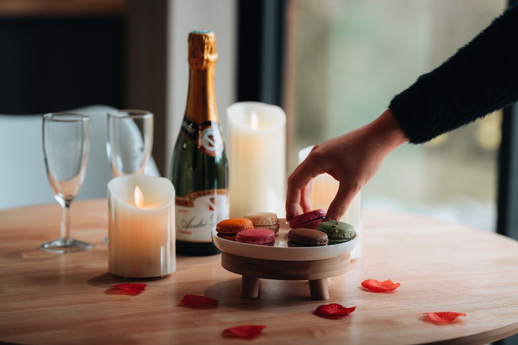 Een kaars, een fles en een bordje met macarons op een houten tafel.