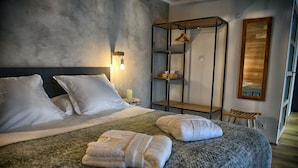 Een net opgemaakt bed met kussens, een grijs gebreid plaid en twee opgevouwen handdoeken. Een metalen wandplank met kledingroede staat naast het bed.