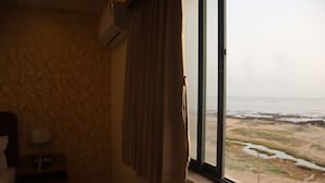 A room with a patterned wallpaper, a window with curtains, a bedside table with a lamp, and a view of the beach. - The Sky Comfort Beach Hotel, Dwarka (Dwarka)