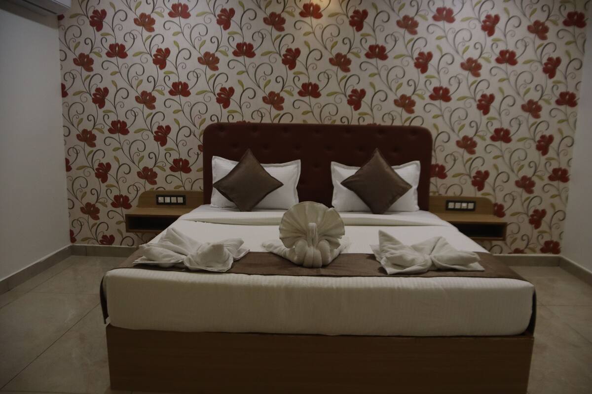 Un lit bien fait, avec une tête de lit marron, du linge de lit blanc et des coussins décoratifs. Il y a deux tables de chevet avec des lampes. Les murs de la pièce sont recouverts d’un papier peint à motifs floraux.