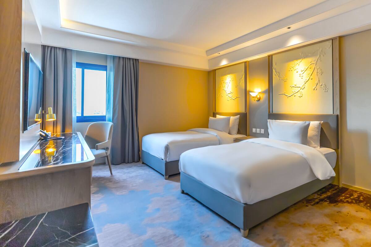 Ein Hotelzimmer mit zwei Betten, einem Schreibtisch, einem Stuhl und einem großen Fenster mit Vorhängen.