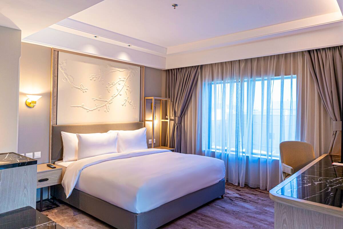 Ein modernes Hotelzimmer mit einem großen Bett, einem Schreibtisch mit Glastischplatte, einem Sessel und Blick auf die Stadt durch große Fenster.