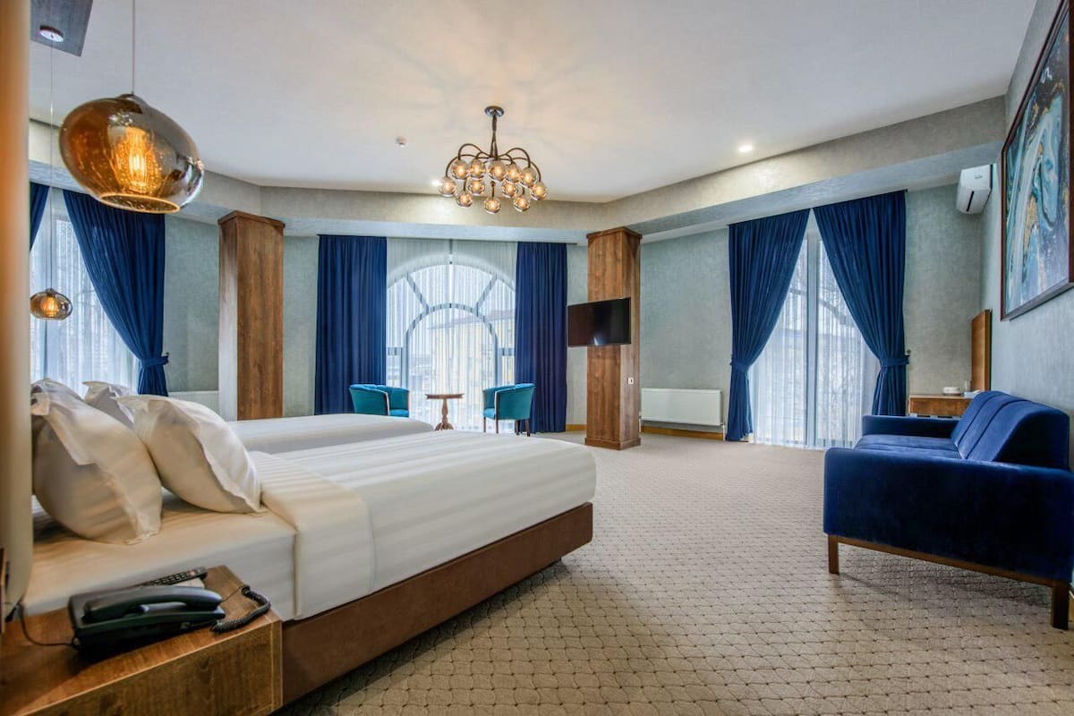 A hotel room with a large bed, a blue sofa, a chandelier, and a view of the city through large windows.