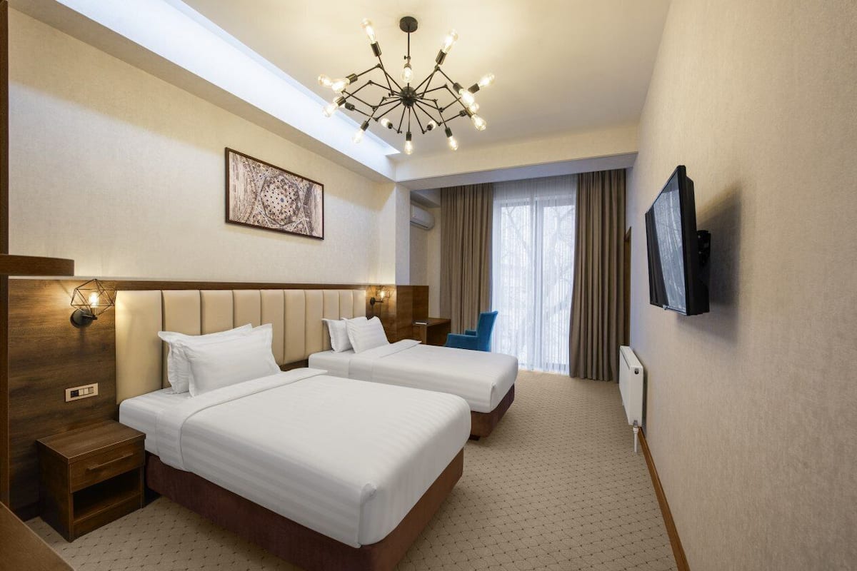 A hotel room with two beds, a modern chandelier, a flat-screen TV, and a window with curtains.