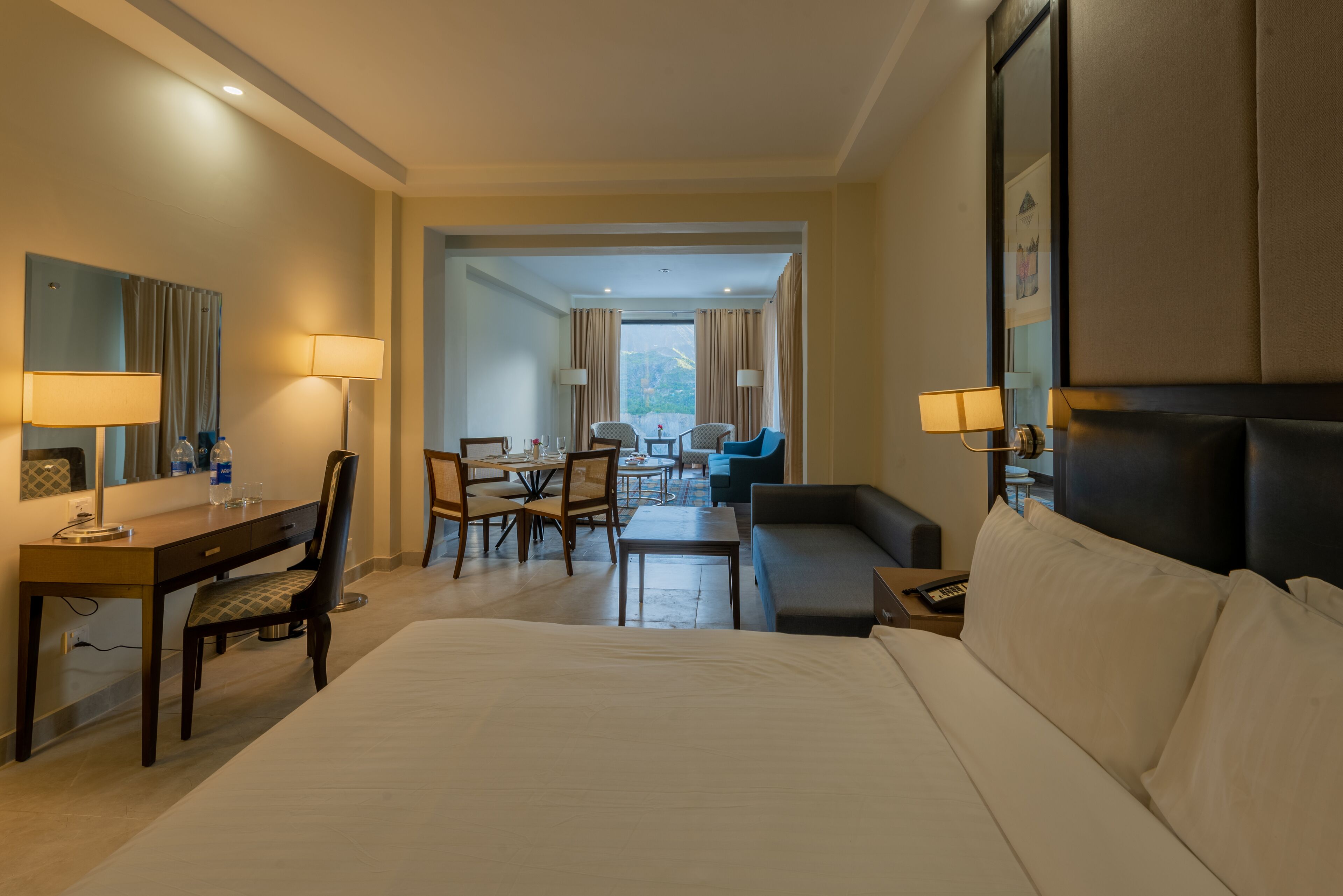 Une chambre d’hôtel avec un grand lit, un bureau avec une chaise, une petite table et un canapé. Il y a deux lampes sur le bureau et un grand miroir au mur.