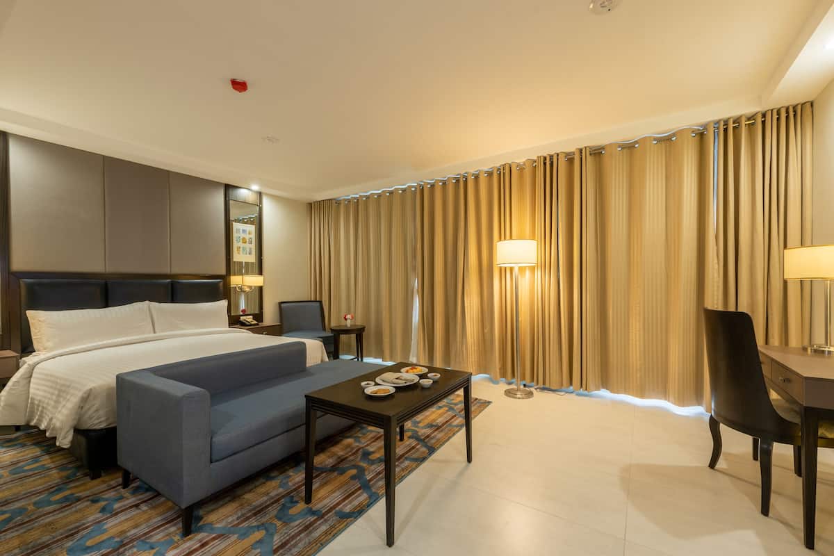 A hotel room with a bed, a sofa, a small table with a lamp, a chair, and a large window with curtains.