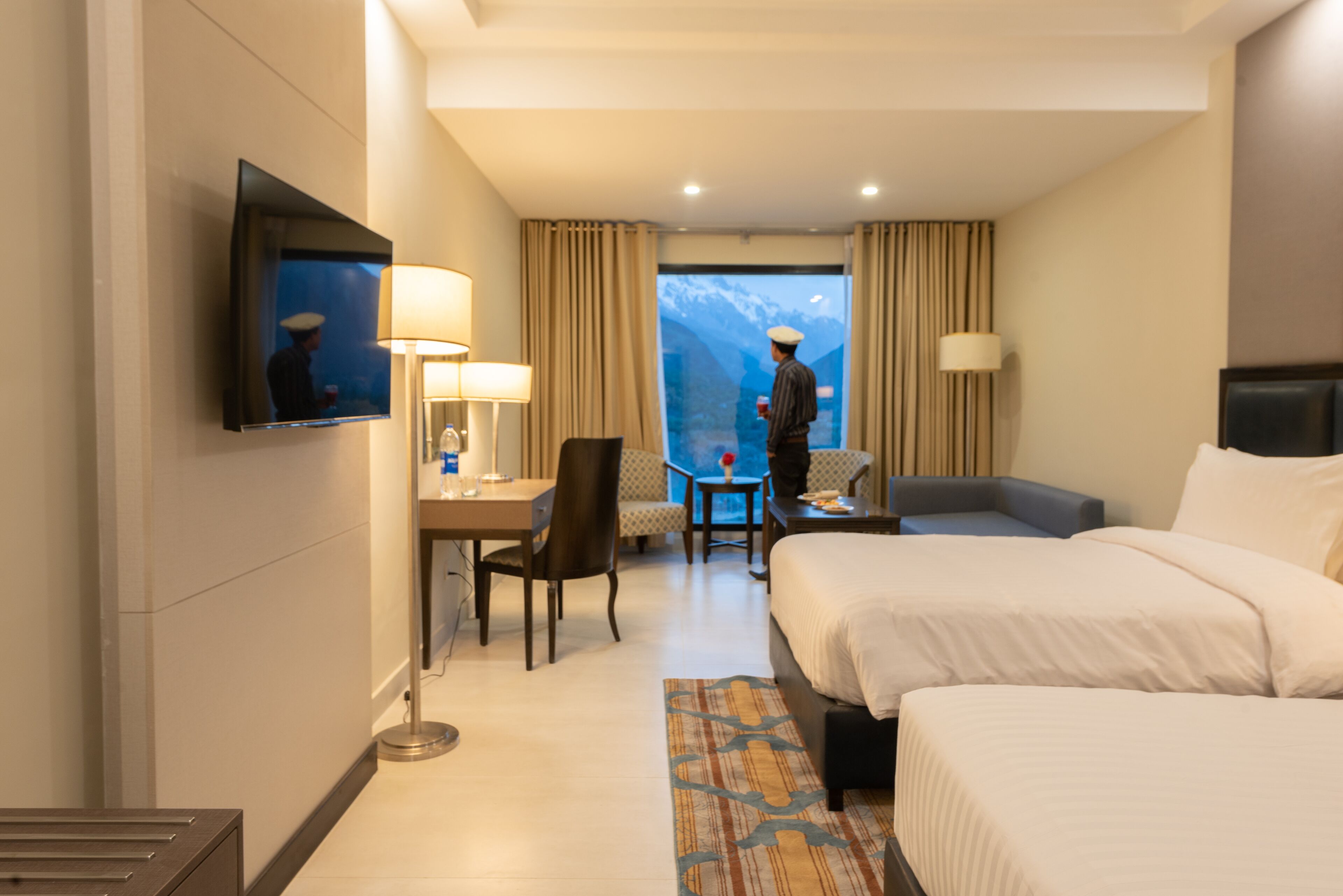 Una habitación de hotel con cama, un escritorio y una silla, un televisor montado en la pared y una ventana con vistas a las montañas.