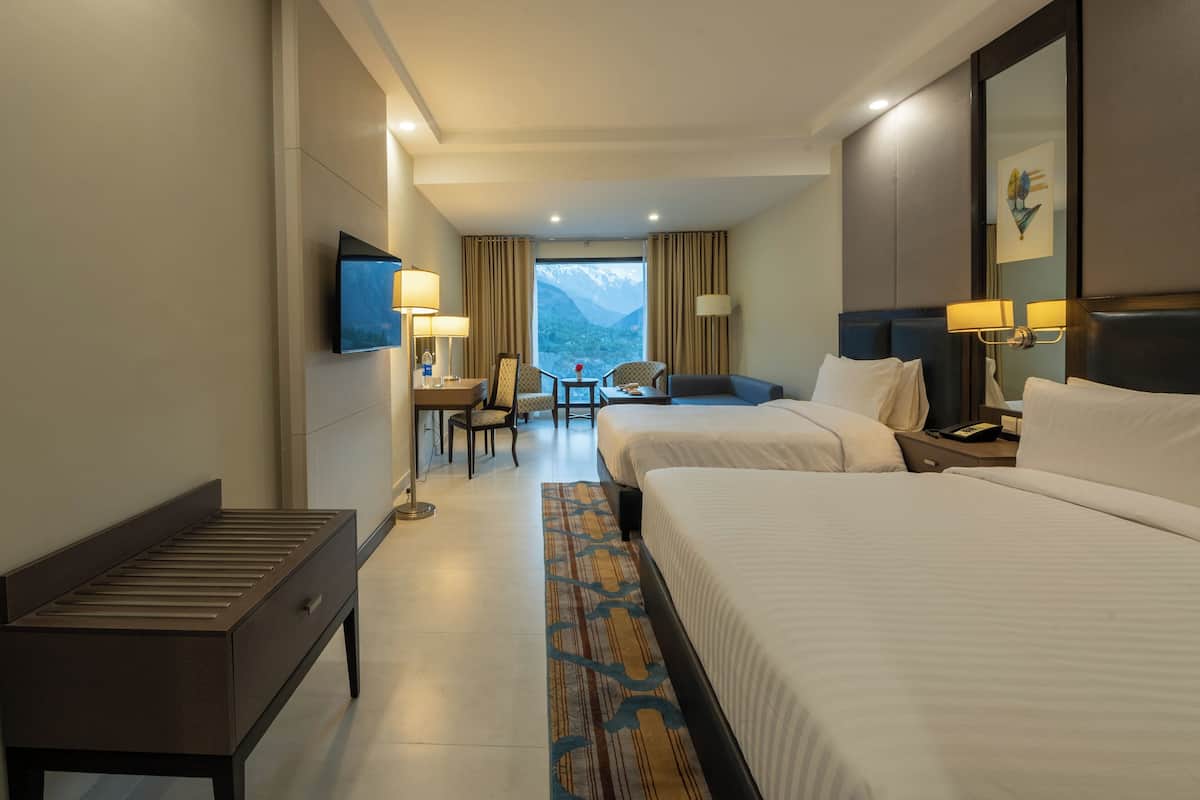 A hotel room with two beds, a desk, chairs, a TV, and a view of mountains.