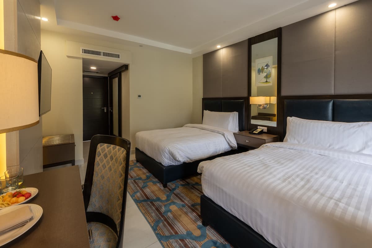 A hotel room with two beds, a desk with a lamp, a chair, and a dining area with plates and glasses.