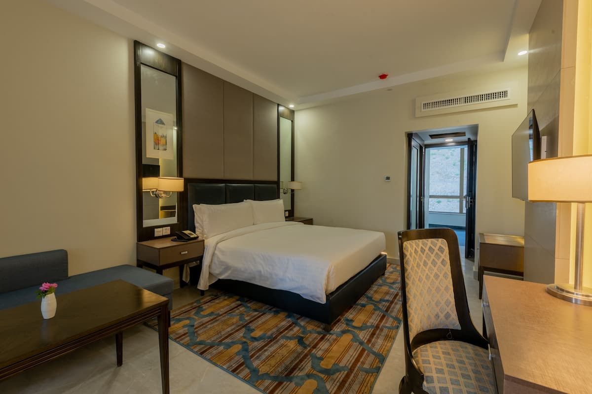 A hotel room with a bed, a desk, a chair, a sofa, a nightstand, a lamp, and a mirror.