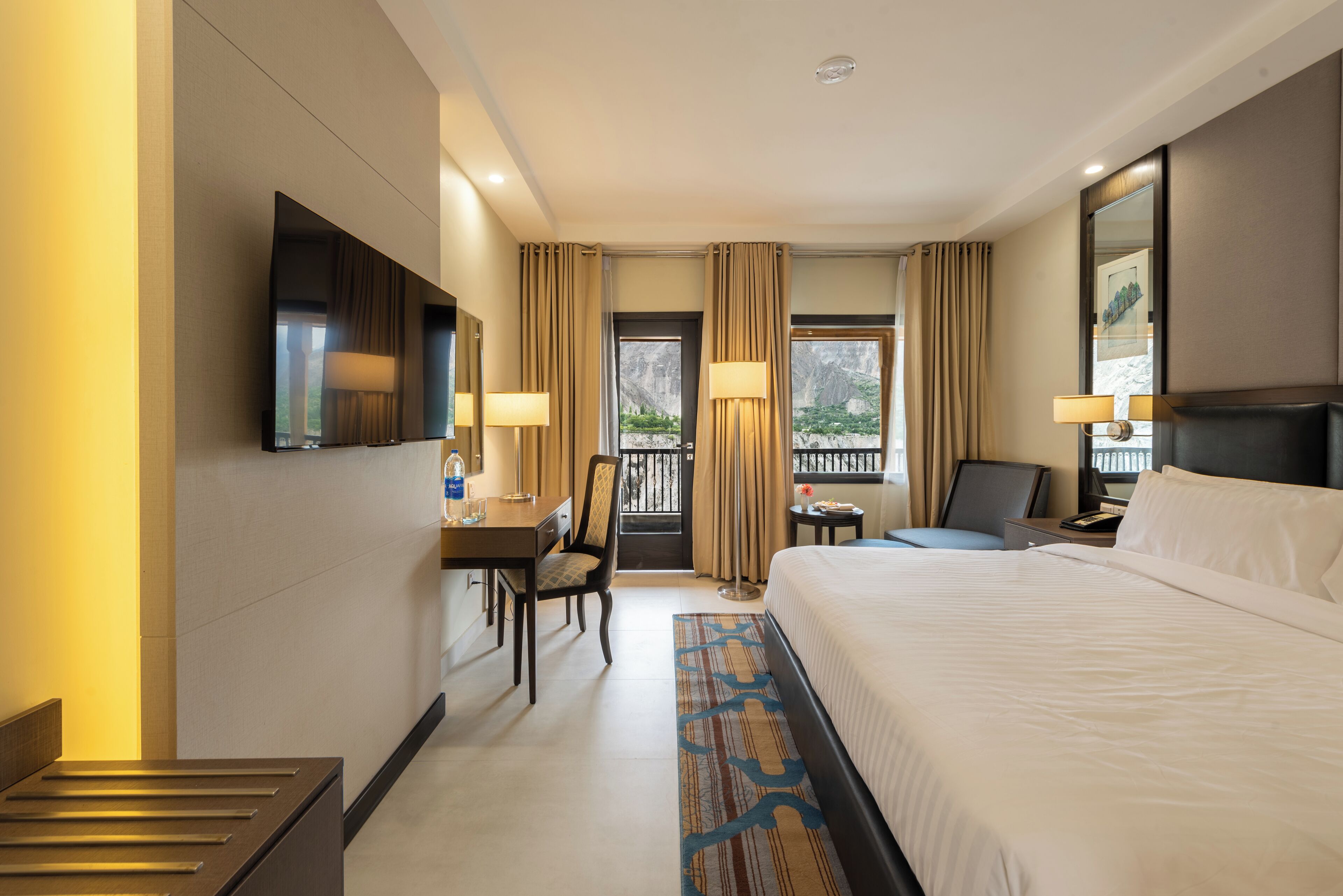 A hotel room with a bed, a desk, a chair, a TV, a window with curtains, and a view of the outdoors.
