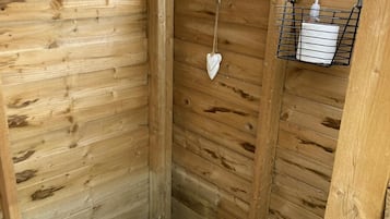 A wooden cabin interior with a wooden wall, a hanging heart-shaped decoration, and a black metal shelf with a tissue box and a bottle.