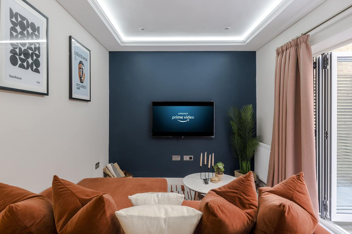 A modern living room with a dark blue accent wall, a mounted flat-screen TV, and a cozy seating area with orange and white cushions.