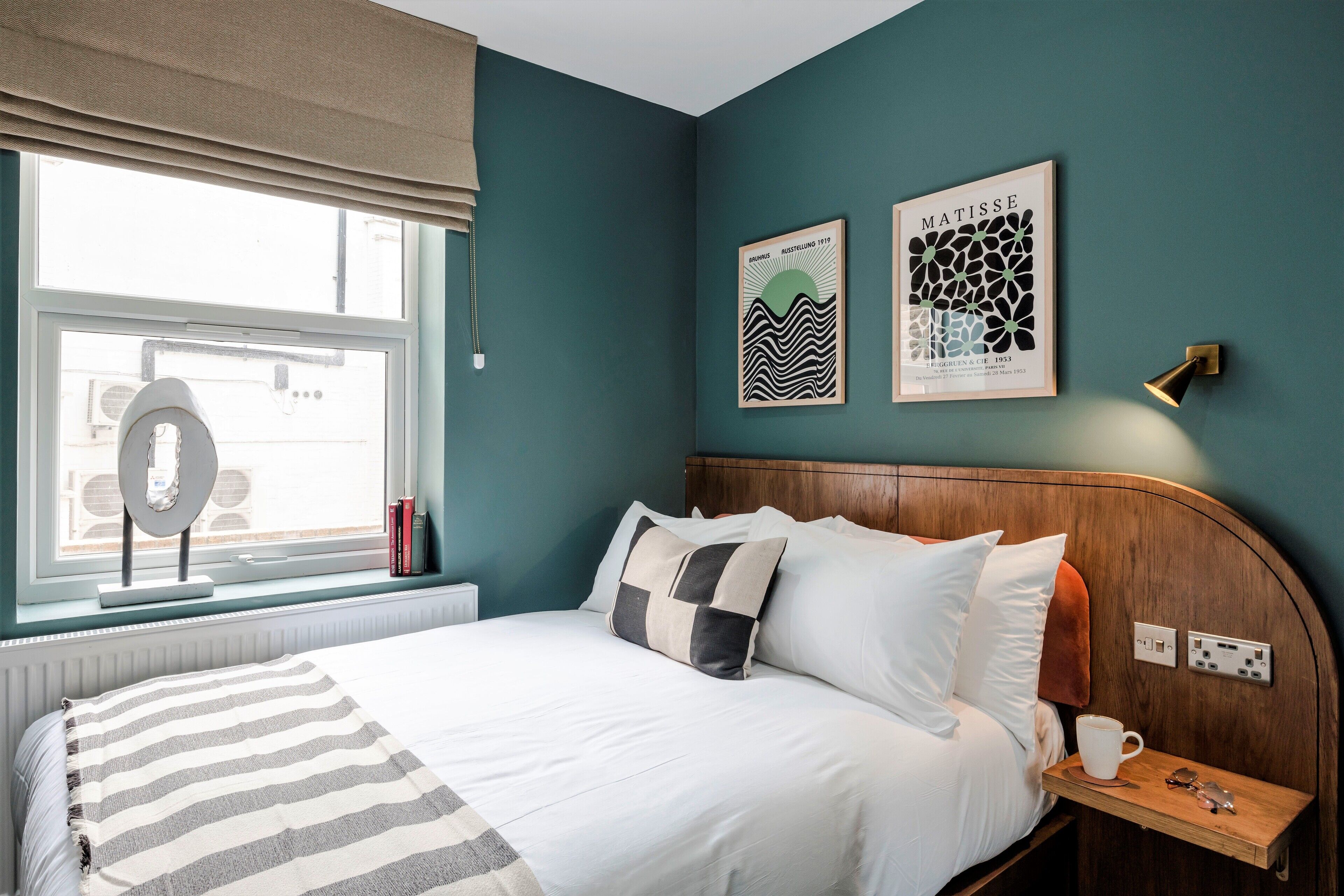 A neatly made bed with a striped blanket, a bedside table with a cup and glasses, and two framed artworks on the wall.