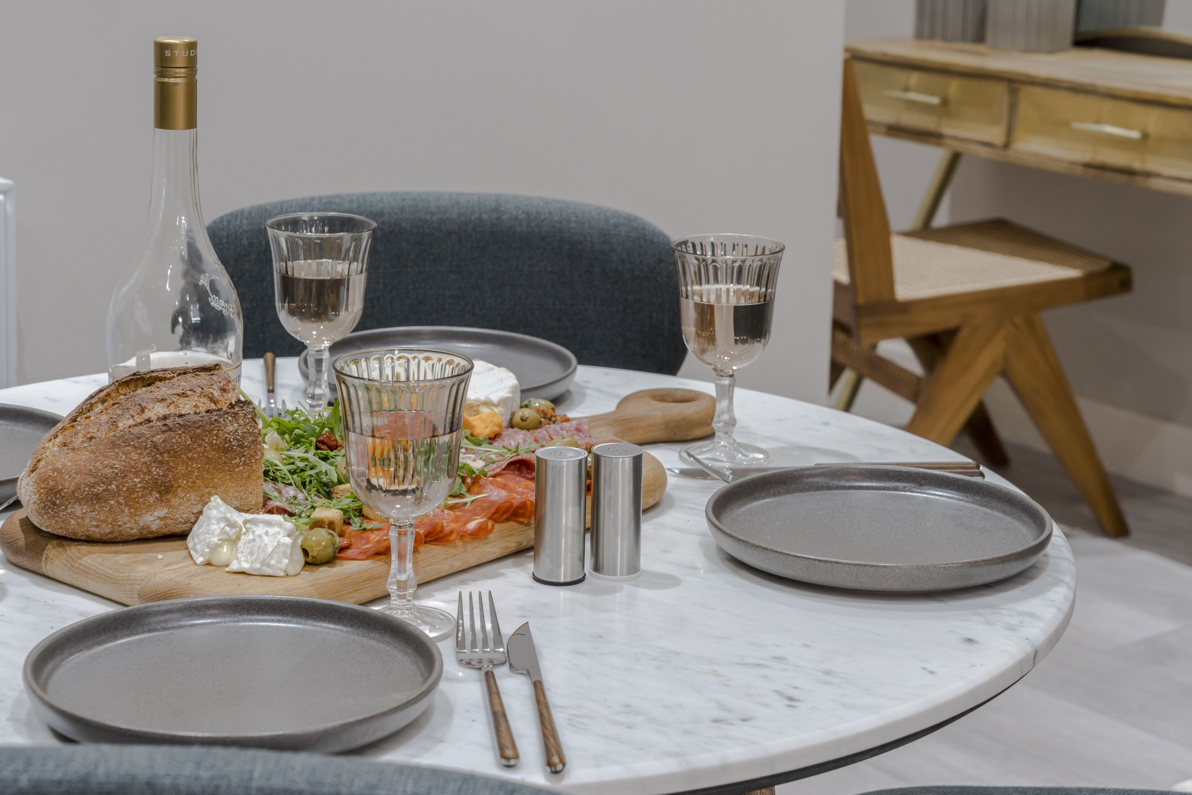 Mesa de comedor puesta con una botella, vasos, platos, cubiertos y una tabla de madera con comida.