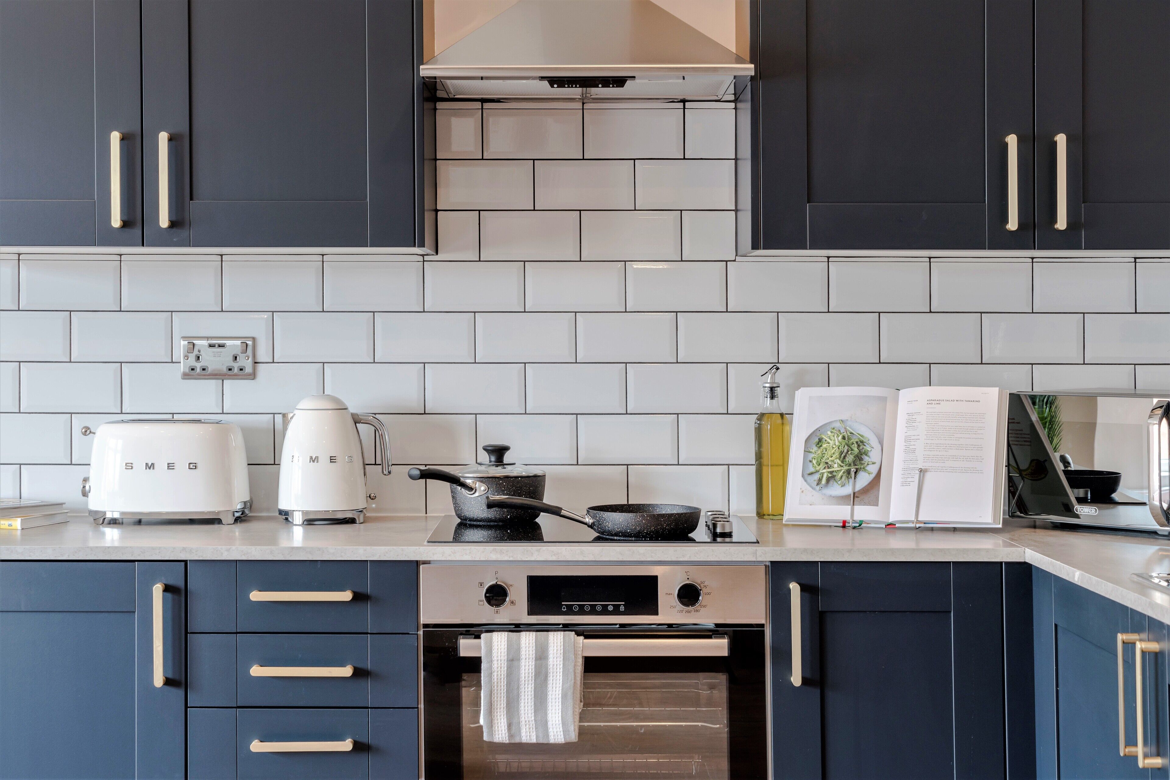 A modern kitchen with dark cabinets, white tiles, a toaster, a coffee maker, a frying pan, and an oven.