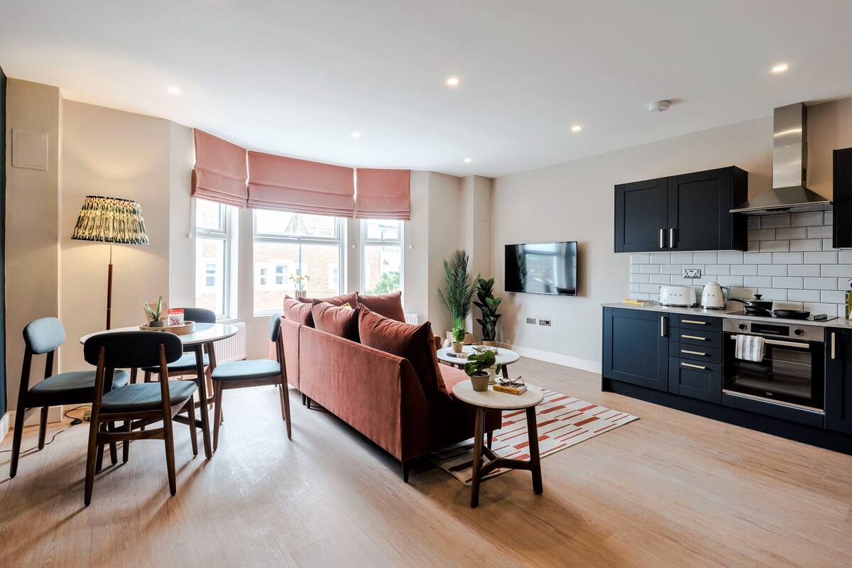 A modern living room with a sofa, chairs, a coffee table, and a lamp. There is a kitchen area with cabinets, a stove, and a TV.