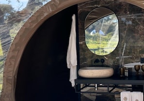 Tienda compacta y circular con un lavabo de piedra y un espejo que refleja un paisaje natural.