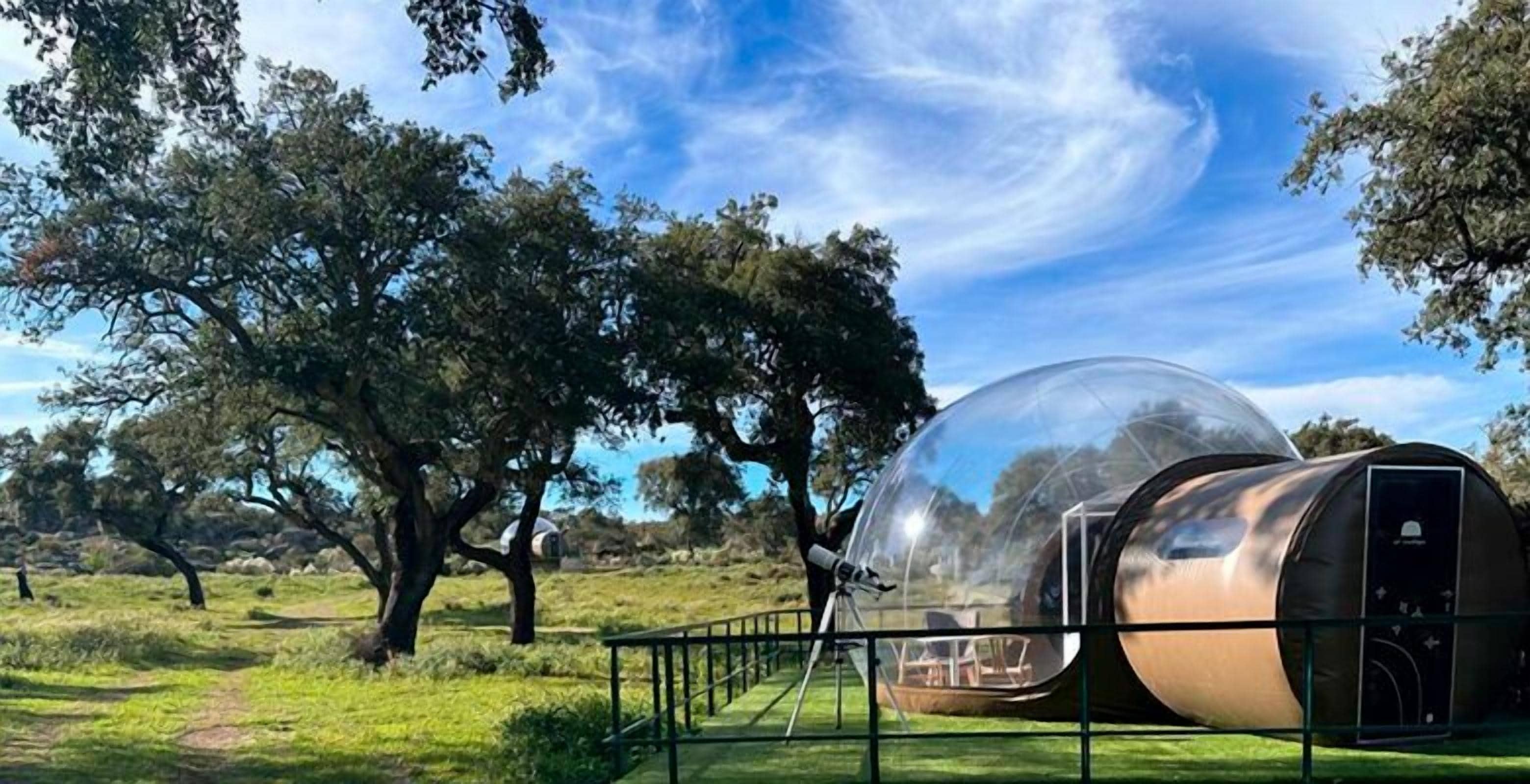 A transparent spherical structure with a metallic exterior, surrounded by a metal railing, set in a grassy field with trees in the background.
