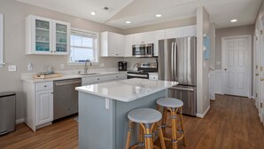 Una cocina moderna con armarios blancos, electrodomésticos de acero inoxidable, una isla grande con taburetes y un ventanal con persianas.