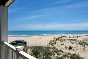 A beach view from a balcony with a car parked outside, sandy beach, and ocean.