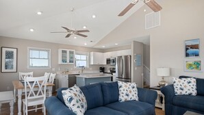 Una sala de estar moderna con un sofá azul, un comedor con sillas blancas, una cocina con armarios blancos y un ventilador de techo.