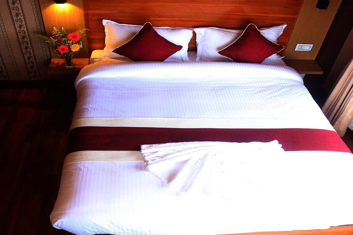 Una cama bien hecha con sábanas blancas y almohadas rojas, un cabecero de madera y una mesita de noche con un jarrón de flores.