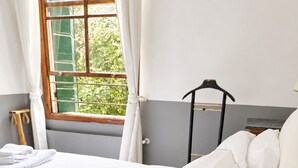 Ein ordentlich bezogenes Bett mit weißen Leinen und Kissen, ein Fenster mit Vorhängen und ein Holzstuhl.