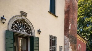 Eine Steinterrasse mit einem runden Holztisch, gedecktem Esstisch, einer Tür mit grünen Fensterläden und einem an der Wand befestigten Laternenaufsatz.