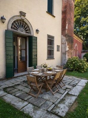 Une terrasse en pierre avec une table ronde en bois, dressée pour le repas, une porte avec des volets verts et une lanterne fixée au mur.