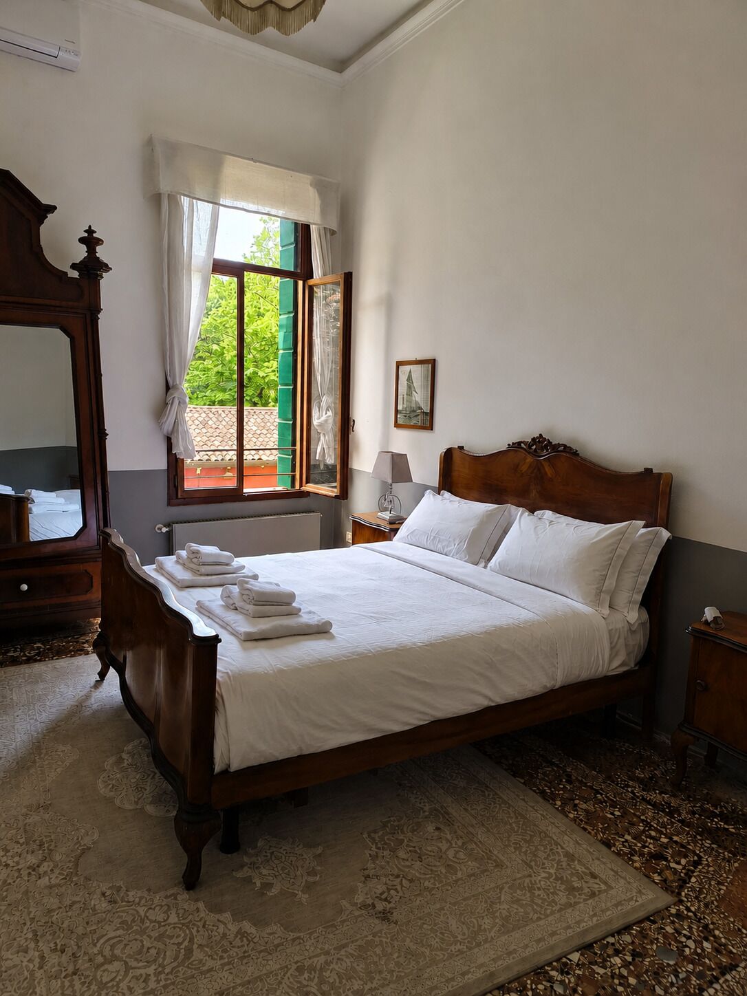 Ein Schlafzimmer mit einem großen Bett, einem Holzschrank, einem Spiegel, einem Fenster mit Vorhängen und Blick ins Grüne.