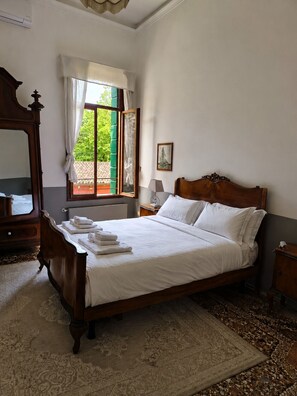 Un dormitorio con una cama grande, un aparador de madera, un espejo, una ventana con cortinas y vistas al verdor exterior.