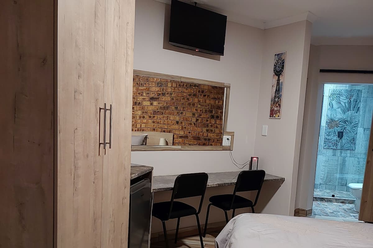 A bedroom with a bed, a desk with two chairs, a TV mounted on the wall, and a window with a brick wall design.
