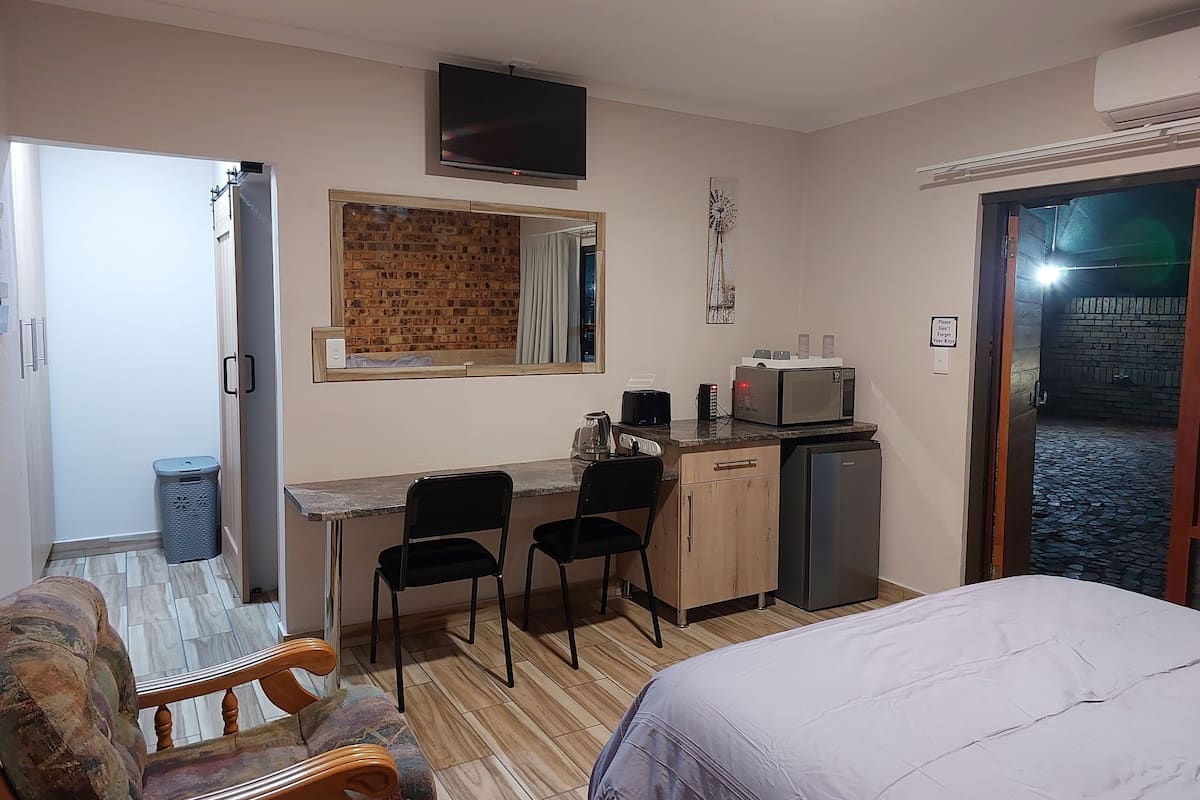 A compact hotel room with a bed, a small kitchenette, a TV, and a brick wall.