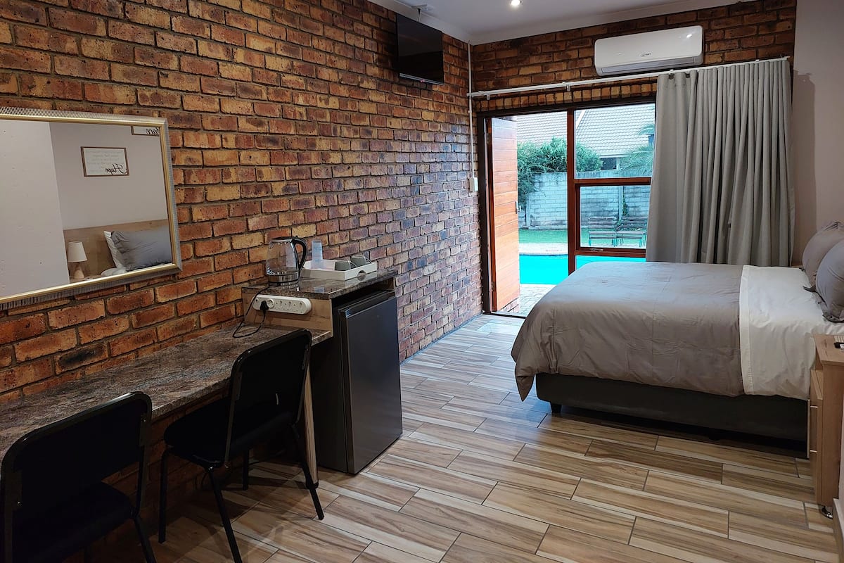 A bedroom with a brick wall, a bed, a desk with a chair, a small kitchenette, and a sliding glass door to a balcony.