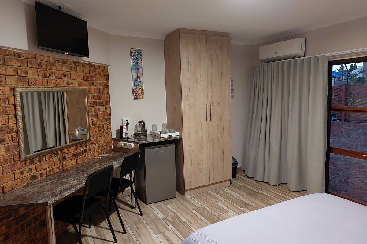 A hotel room with a brick wall, a desk with a mirror, a small fridge, a bed, and a window with curtains.