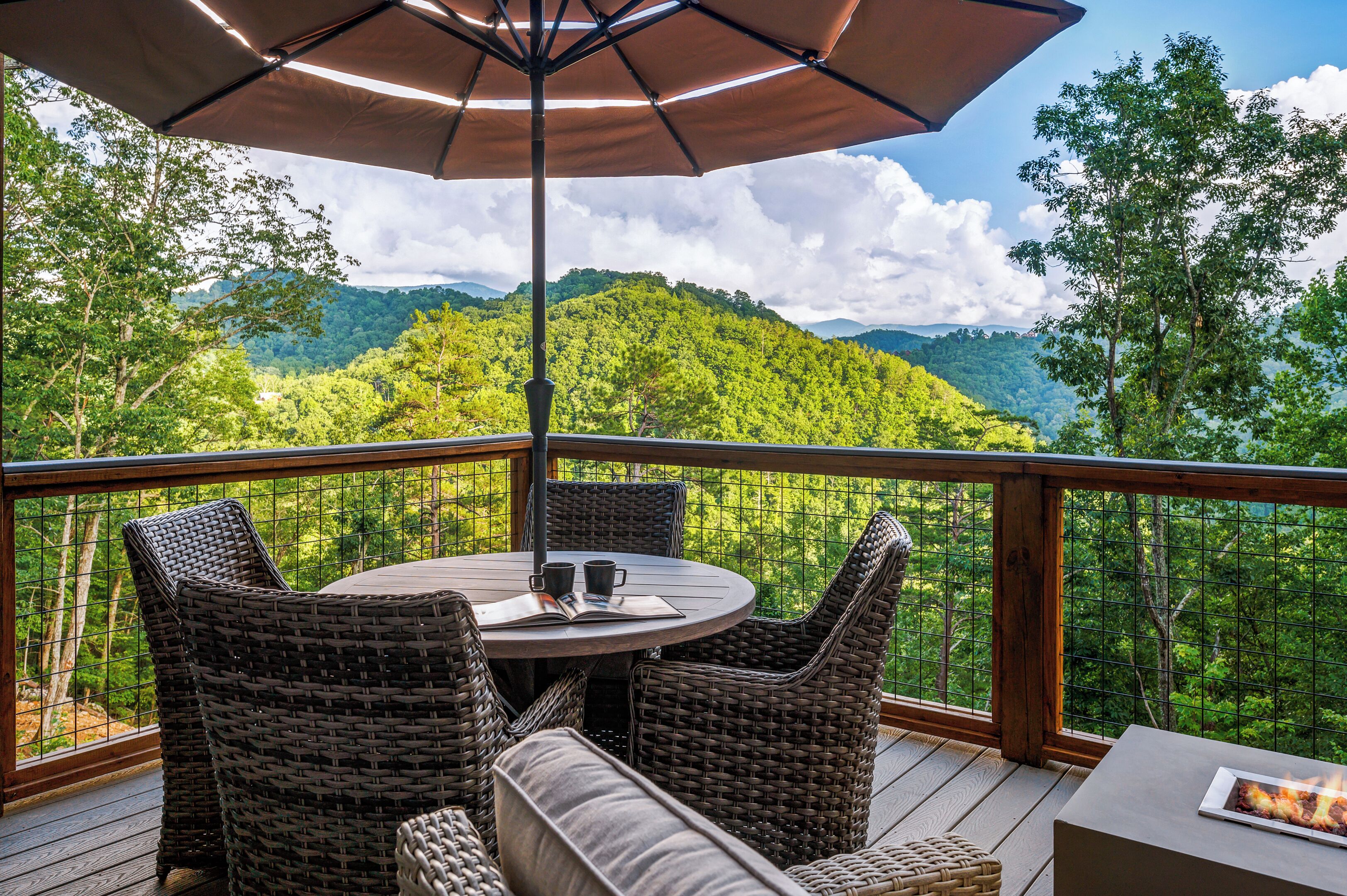 A deck with wicker furniture, a table, and an umbrella, overlooking a forested mountain landscape.