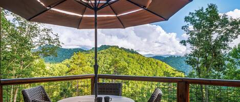A deck with wicker furniture, a table, and an umbrella, overlooking a forested mountain landscape.