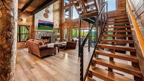 A spacious living area with a stone fireplace, wooden staircase, and large windows offering views of the outdoors.