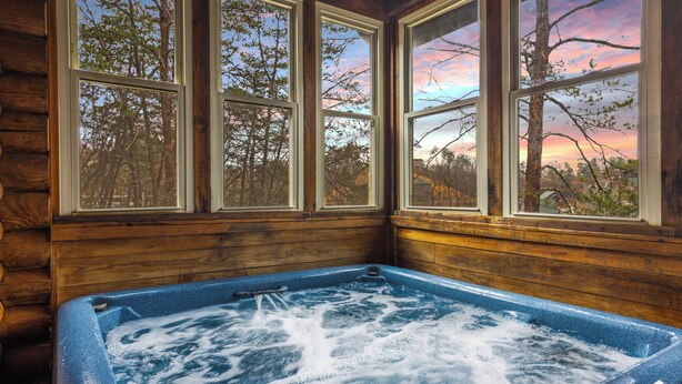 A wooden cabin interior with a hot tub and large windows overlooking a forested area.