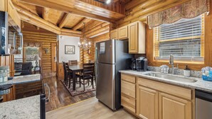A log cabin interior with a kitchen featuring wooden cabinets, a stainless steel refrigerator, and a large window with blinds.