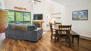 Ein Wohnzimmer mit grauem Sofa, einem Holzesstisch, Stühlen, einem Kamin und einem an der Wand befestigten Fernseher.