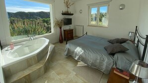 Une chambre avec un grand lit, une table de chevet, une commode, une télévision, une fenêtre donnant sur un paysage verdoyant et une baignoire.